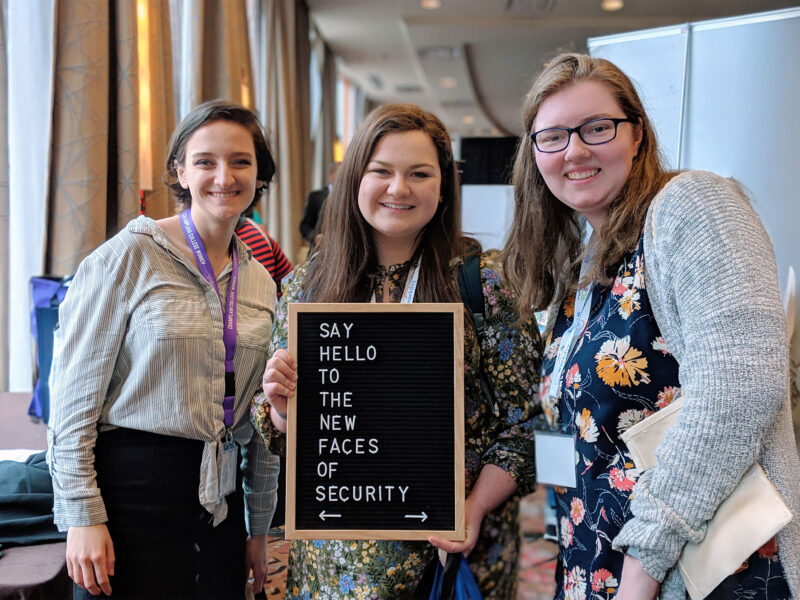 Students at a conference hold a sign that says "Say hello to the new faces of cybersecurity."
