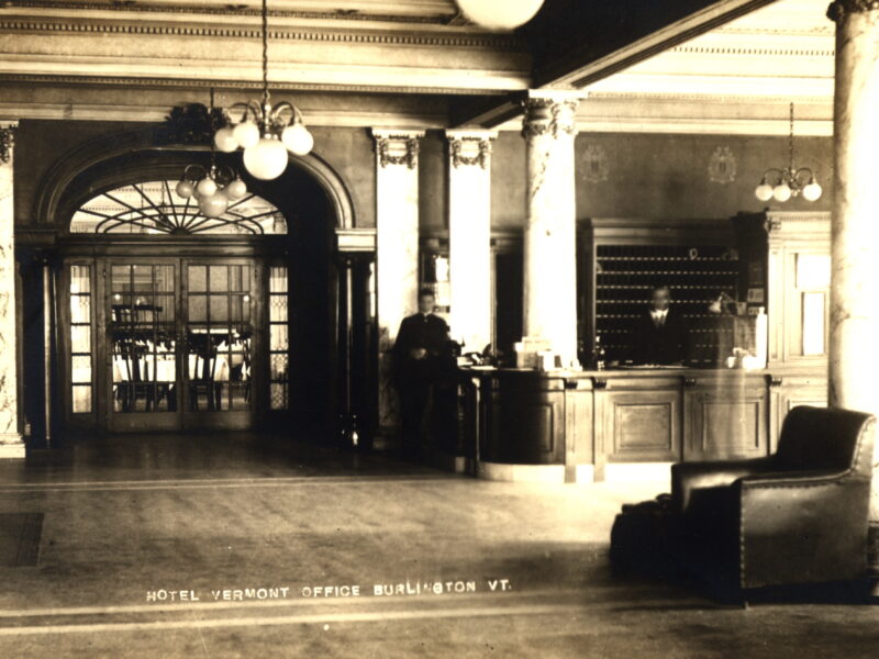 Black and white photo postcard (circa 1910) of the Hotel Vermont lobby in Burlington, VT, showing the front desk, ornate columns, and dining room in the background.