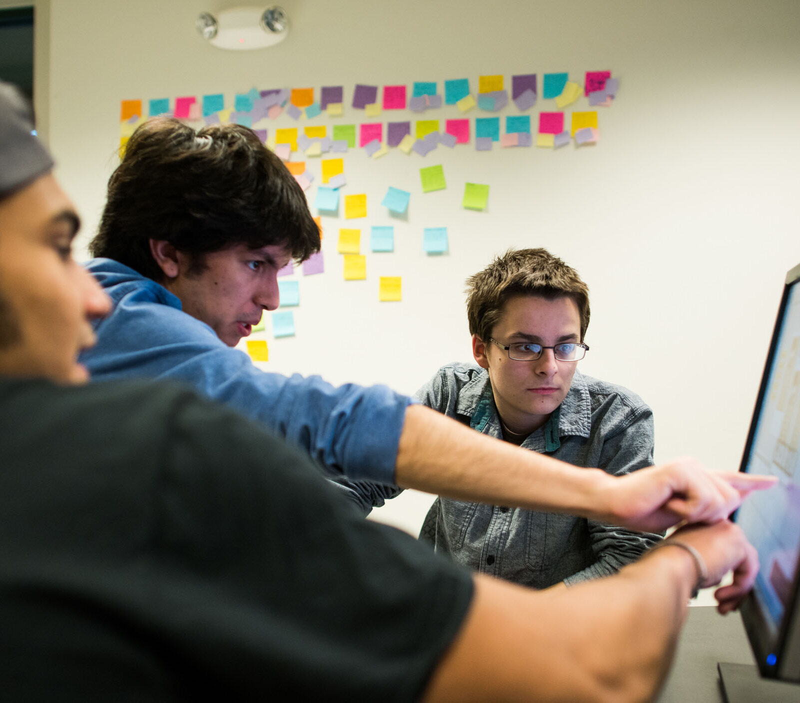 three students working on a computer in front of a wall of post-it notes