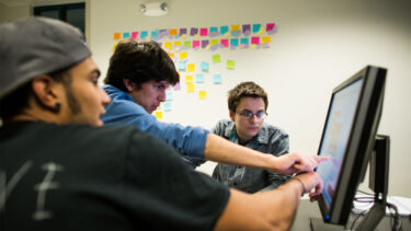 three students working on a computer in front of a wall of post-it notes