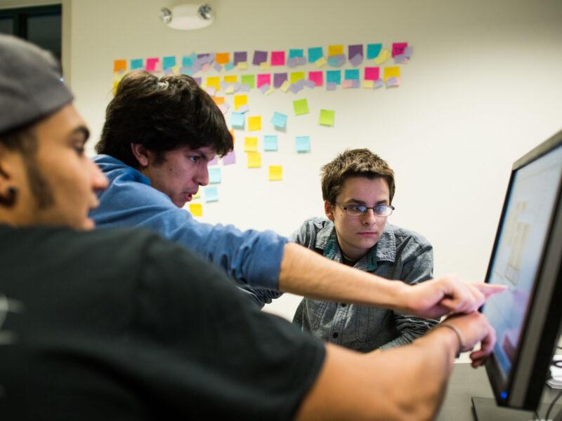 three students working on a computer in front of a wall of post-it notes