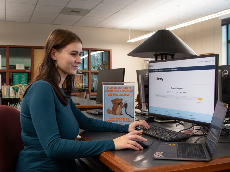 A smiling student uses a laptop connected to a USB-C docking station monitor displaying the library's Research Navigator website at a library workstation.