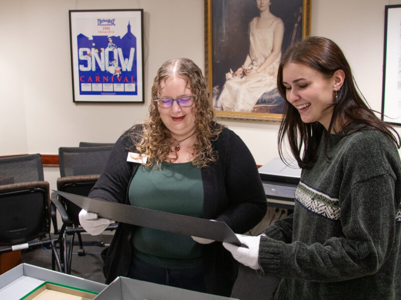 A staff member and student wearing white conservation gloves examine an archival document together and smile in a special collections reading room.