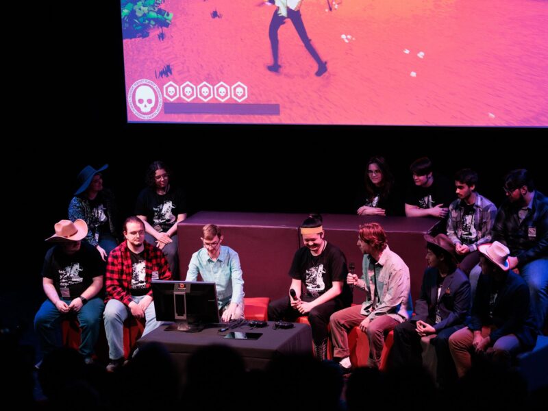 A group of students sit together in front of a screen, presenting their game.
