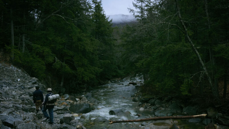 Two people walking in the wilderness near a river with a mountain range in the background, surrounded by trees