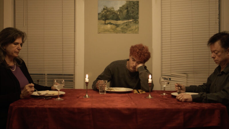 three people sitting at a red table in a house, the person in the middle slouched over while the others sit upright