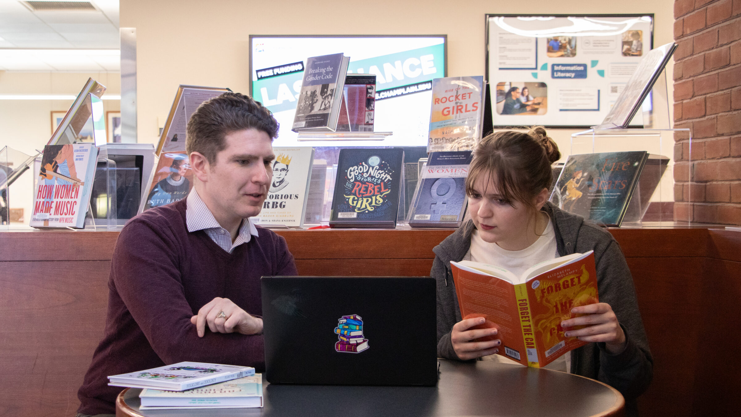 A librarian and student sit together at a table in the library, the student reading a book while the librarian explains to them the research on the laptop in front of them, with a book display in the background.