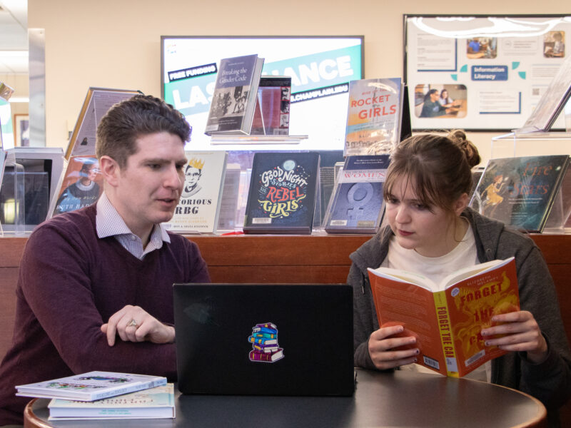 A librarian and student sit together at a table in the library, the student reading a book while the librarian explains to them the research on the laptop in front of them, with a book display in the background.