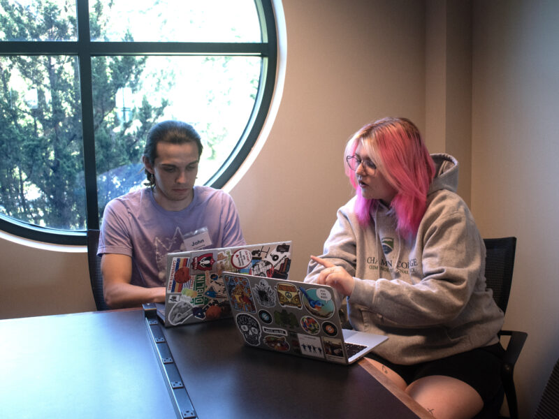Two students collaborate on laptops at a table in the Miller Information Commons.
