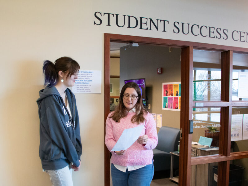 A student and advisor meet outside the entrance to the Student Success Center, the advisor explaining paperwork during their conversation.