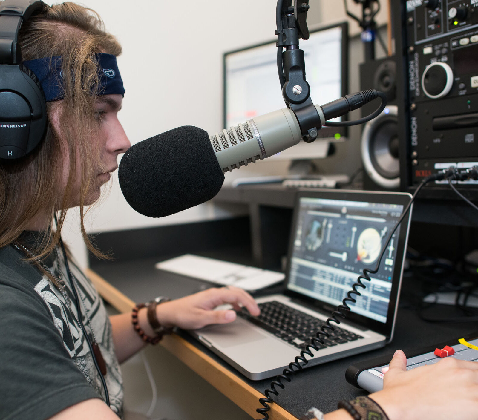 A student wearing a headset while leaning into a mic, using a laptop and sound recording equipment