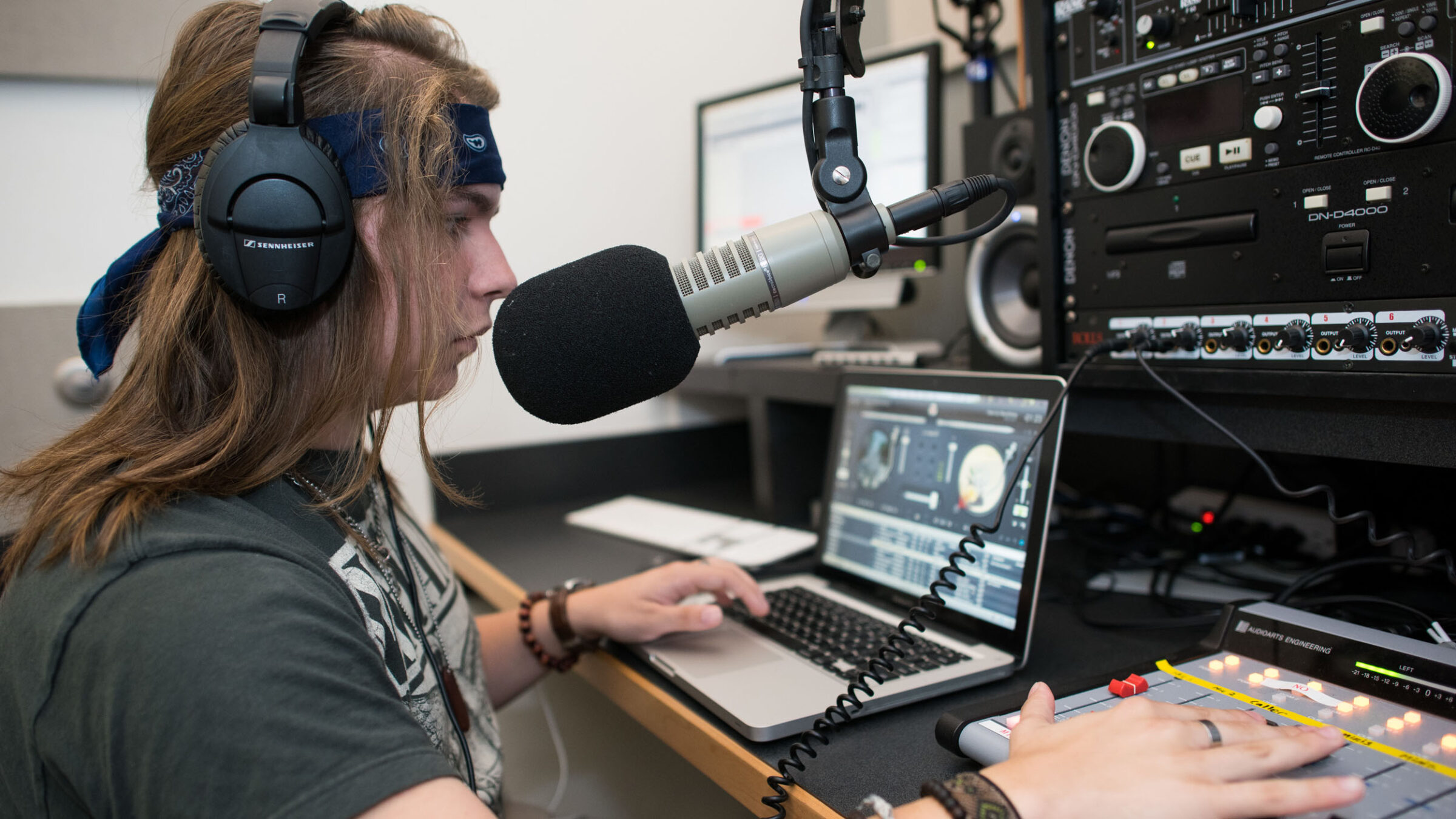 A student wearing a headset while leaning into a mic, using a laptop and sound recording equipment