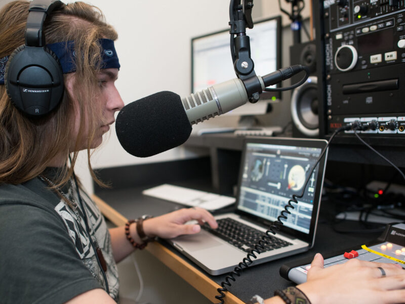 A student wearing a headset while leaning into a mic, using a laptop and sound recording equipment