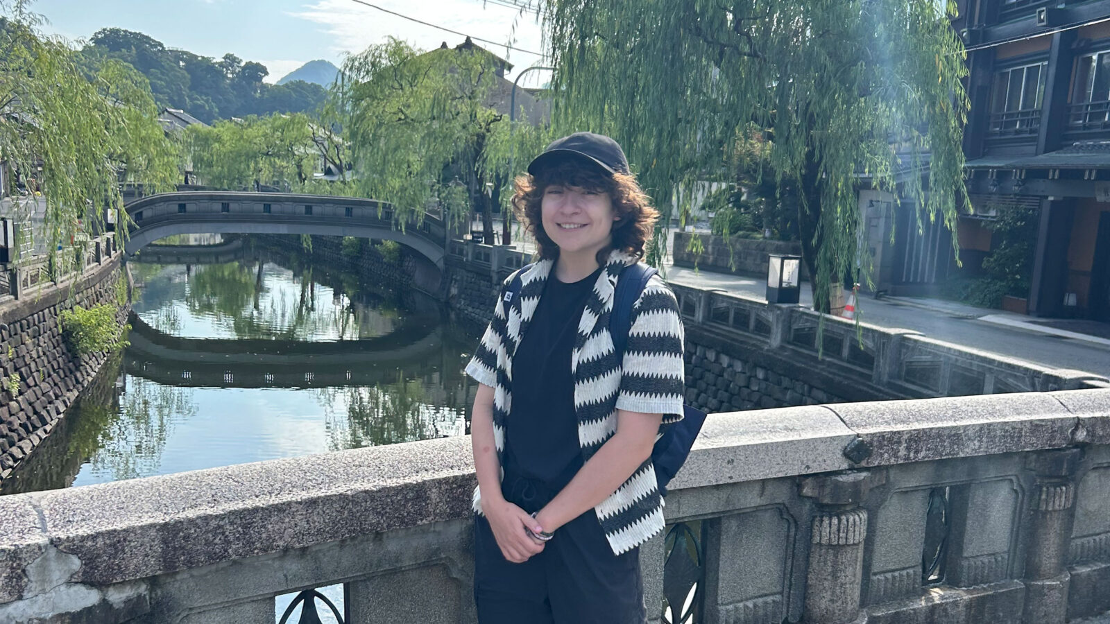 student stands smiling on a bridge with greenery behind them