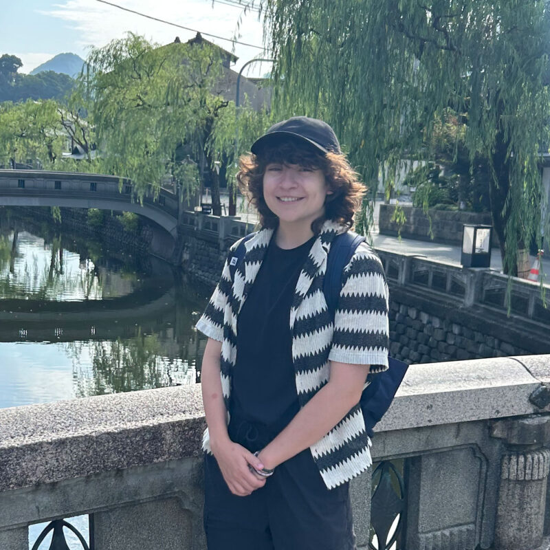 student stands smiling on a bridge with greenery behind them