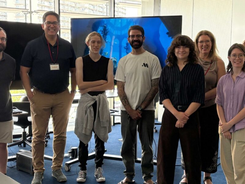 a group of students and President Alex Hernandez and his wife Michelle pose and smile at the camera with a big TV screen in the background