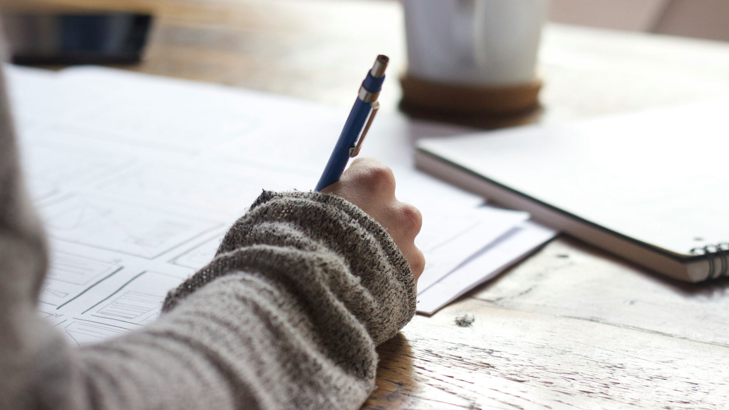 A person sitting at a desk with paper, a pen, and a mug writing.