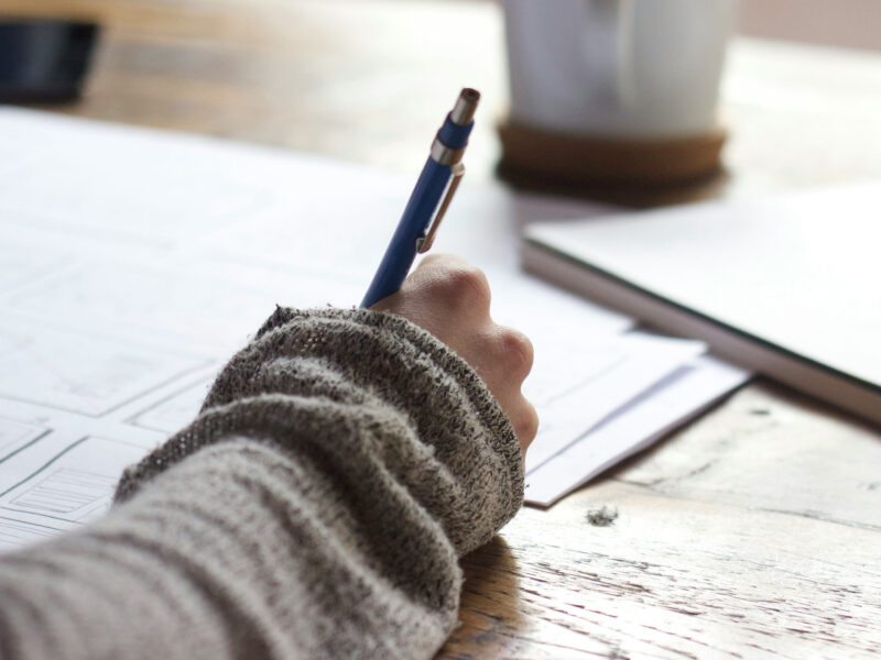 A person sitting at a desk with paper, a pen, and a mug writing.