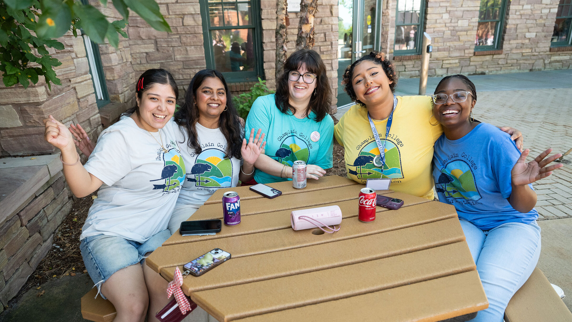 five students sit at a picnic table smiling at the camera with their hands in the air