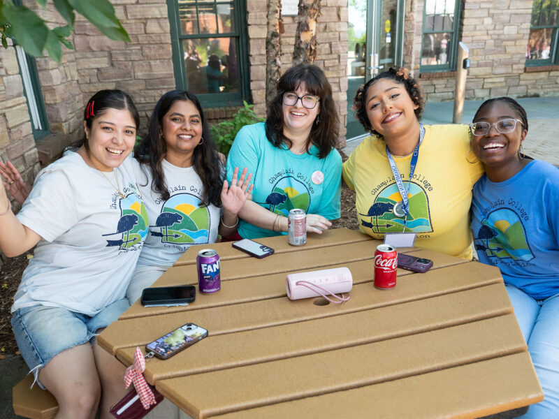five students sit at a picnic table smiling at the camera with their hands in the air