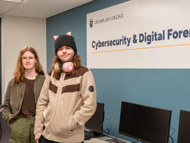 two students pose for the camera standing in front of a "champlain college cybersecurity & digital forensics" sign