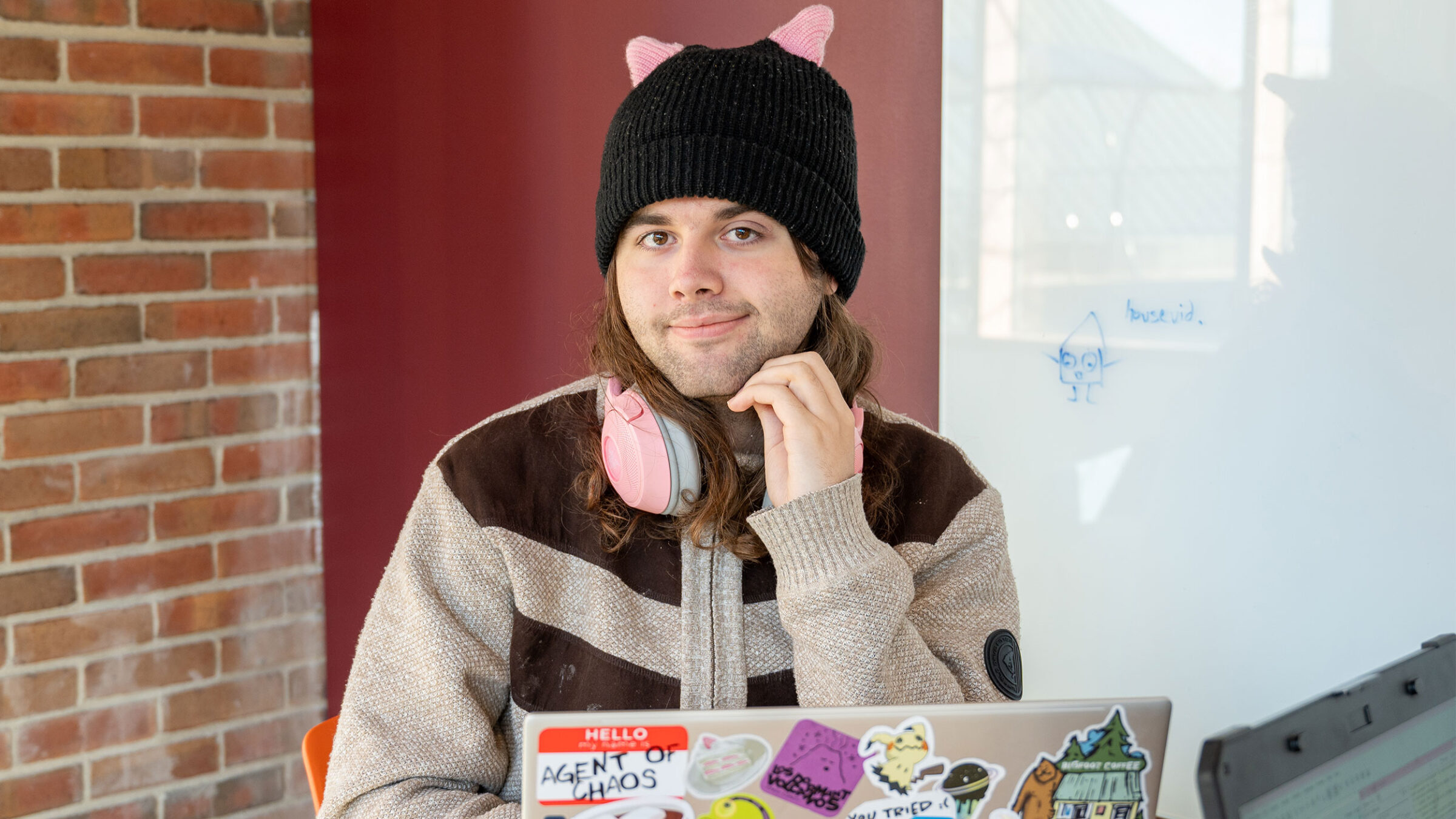 student wearing tan and black sweater and a pink and black hat smiles while working on a laptop