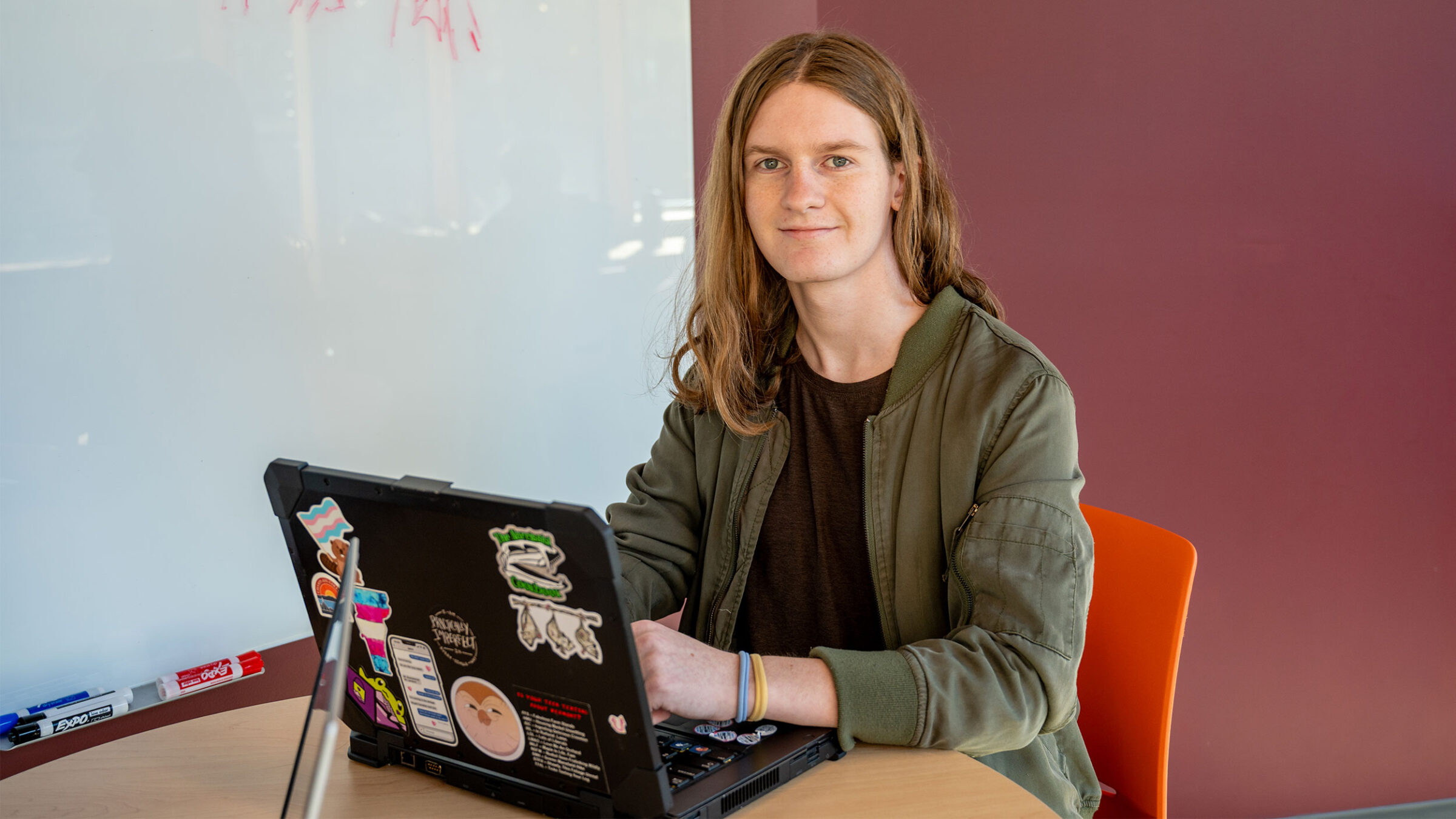 student in green jacket and black t-shirt smiles at camera while working on a laptop
