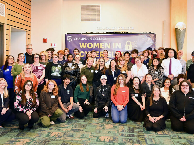 large group of women in cyber attendees pose for photo in front of a "women in cyber summit" blue sign