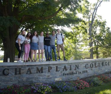 A group of students gathered together on top of a stone wall with the Champlain College welcome sign.