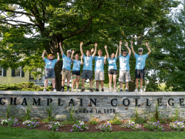 Orientation Leaders waive while standing on a stone wall Champlain College sign